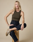 A woman with shoulder-length blonde hair wears Sold Out NYC's best-selling "The Not So Basic Tank" in dark olive, paired with dark high-waisted jeans and beige sneakers, as she sits on a wooden stool against a plain beige backdrop.