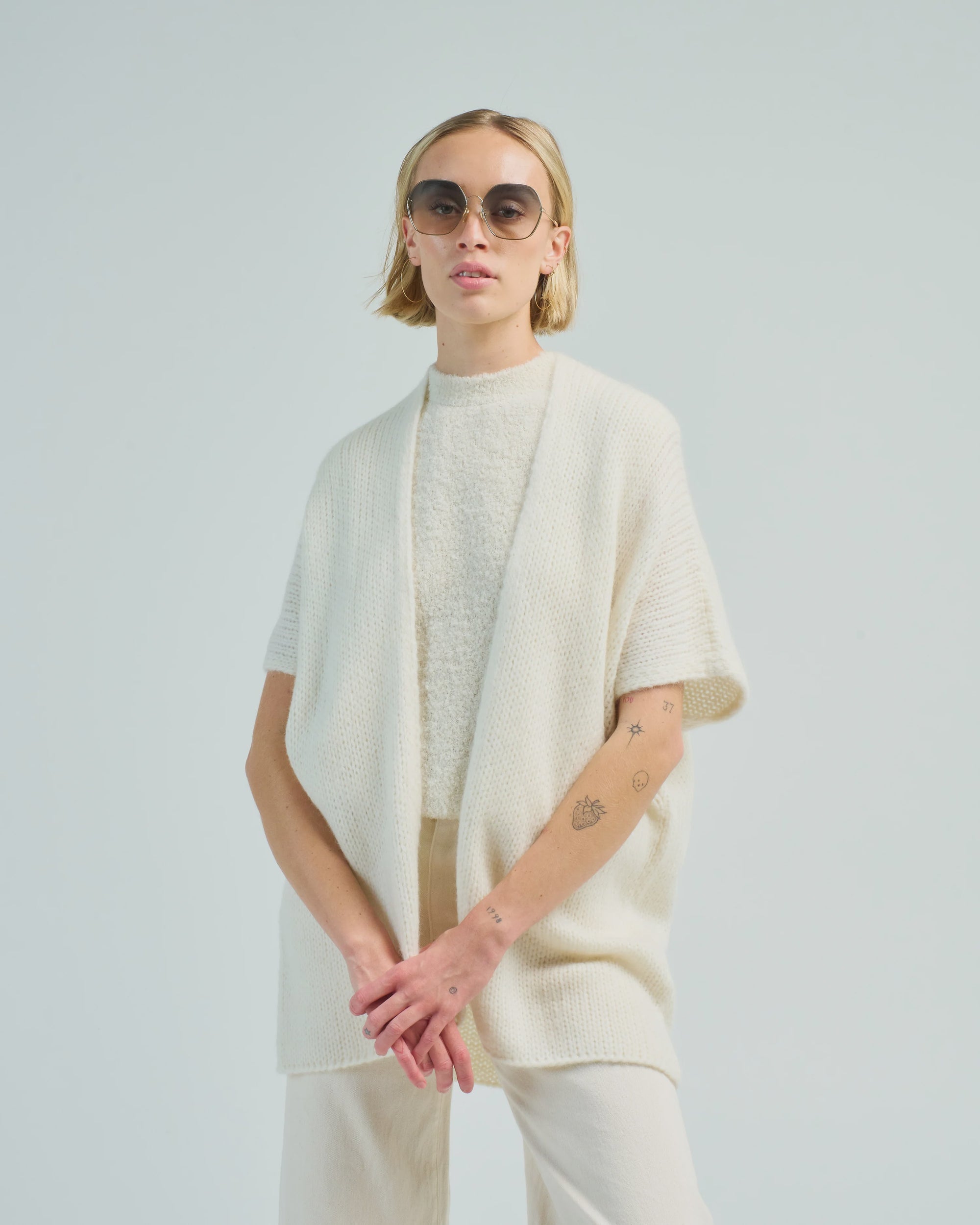 A person with short blonde hair, round sunglasses, a textured white top, the LaTierra Nube Vest, a loose cream cardigan, and light pants stands against a plain light background with arms loosely crossed.
