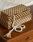 Two Made Goods Zion Hair on Hide Boxes, featuring a geometric gold and black diamond pattern, sit atop a wooden surface. A string of white beads with a tassel is draped over them, enhancing their sophisticated style.