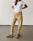 A person stands indoors against a plain wall, wearing a white t-shirt, Hey Gang’s The Officer Pant in cotton ripstop with rolled cuffs, and light brown slip-on shoes. Their hands are on their hips, and their face is out of the frame.