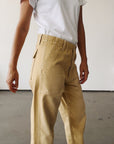 A person stands indoors against a plain white wall, arms at their sides, wearing a white t-shirt and Hey Gang’s The Officer Pant—100% Cotton Ripstop, high-waisted tan work pants with a relaxed leg.