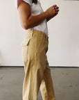 A person stands indoors against a plain white wall, hands clasped near their chest, wearing Hey Gang’s The Officer Pant and a loose white cropped short-sleeve shirt.