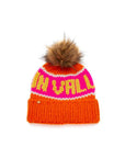 The Faire Knit Ski Hat is a luxury merino wool beanie with a brown faux fur pom-pom, an orange cuff, a pink top, and yellow text.