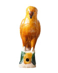 The Parrot Small by Blue Ocean Traders is a ceramic sculpture inspired by Jingdezhen Hand-Painted Porcelain, glazed in yellow and brown, perched on a green and white base with a hole near the bottom, set against a white background.