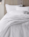 A cozy bed with fluffy white linens and pillows features the Coyuchi Inc Org Relaxed Linen Duvet in Alpine White, crafted from organic linen and low-impact dyes, set against a light wooden headboard beside a dark round bedside table.