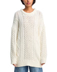 A person wears the Denimist OVERSIZED ARAN SWEATER—a loose, chunky-knit cream cotton piece with cable patterns and ribbed details—paired with blue jeans. The cropped image emphasizes the sweater’s cozy, oversized fit.