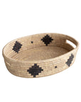 The Faire Palm Leaf Oval Basket is a shallow, handwoven tray with two handles, featuring a natural tan color and bold black geometric patterns—expertly crafted by fair trade artisans.