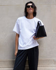 A person with short, wet hair and sunglasses stands against a beige stone wall, wearing The Oversized Tee by Sold Out NYC with black pants and holding a black handbag under one arm.