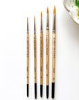 Five Faire Paintbrush Set round brushes, sizes 0, 2, 4, 6, and 8 with synthetic bristles and lacquered wood handles, are lined up on a white surface. A watercolor ceramic palette is partly visible in the top right corner.