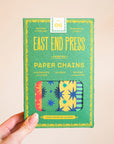 A hand holds a box labeled "Faire Paper Garland/Chains," featuring colorful star and geometric designs—ideal for vibrant decorations or festive birthday garlands. The green packaging displays yellow, blue, and orange text.
