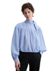 A person wears the Rachel Comey Parachute Top—a wrinkle-resistant, light blue, long-sleeve blouse with a tie collar and loose fit—paired with black pants, hands in pockets against a white background; ideal for travel.