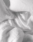 A close-up of Coyuchi Inc’s Org Relaxed Linen Duvet in Alpine White (Queen) with white organic linen sheets and pillows, softly rumpled and unmade, showcasing breathable fabric and low-impact dyed finish.