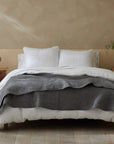 A neatly made bed with white pillows, a white sheet, and the Coyuchi Inc Pebbled Org Quilt in Queen Slate adds texture. Wooden nightstands hold a sculpture, potted plant, and books. Light wood floors and beige walls complete the serene room.