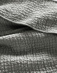 Close-up of the Pebbled Org Quilt Queen Slate by Coyuchi Inc, featuring textured, gray organic cotton with visible stitching and a soft, crinkled look, folded and layered on a surface.