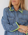A woman stands with folded arms, her face partially visible, wearing the Kerri Rosenthal Pia Denim Western Shirt featuring bright yellow-green trim on the collar and cuffs, and light-colored rings on her fingers.