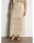 A person wearing the SUNCOO Fanny Skirt in beige crochet with floral patterns and lace detailing, paired with black heeled sandals and a silver bracelet, stands against a white textured wall.