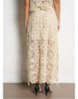 A person stands against a white textured wall wearing the SUNCOO Fanny Skirt in cream, featuring intricate lace and floral patterns. The semi-sheer skirt is elegantly paired with black high-heeled sandals.