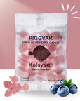 A bag of Faire Piggvar Candy Fish with a transparent window is shown, surrounded by pink gummy candies and fresh blueberries. This vegan, gluten-free treat honors classic Swedish confectioneries.