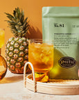 A glass and pitcher of iced tea sit on a tray with peach, pineapple, and mango, next to a green package labeled "Smith Tea" by Faire, all set against a warm yellow background.