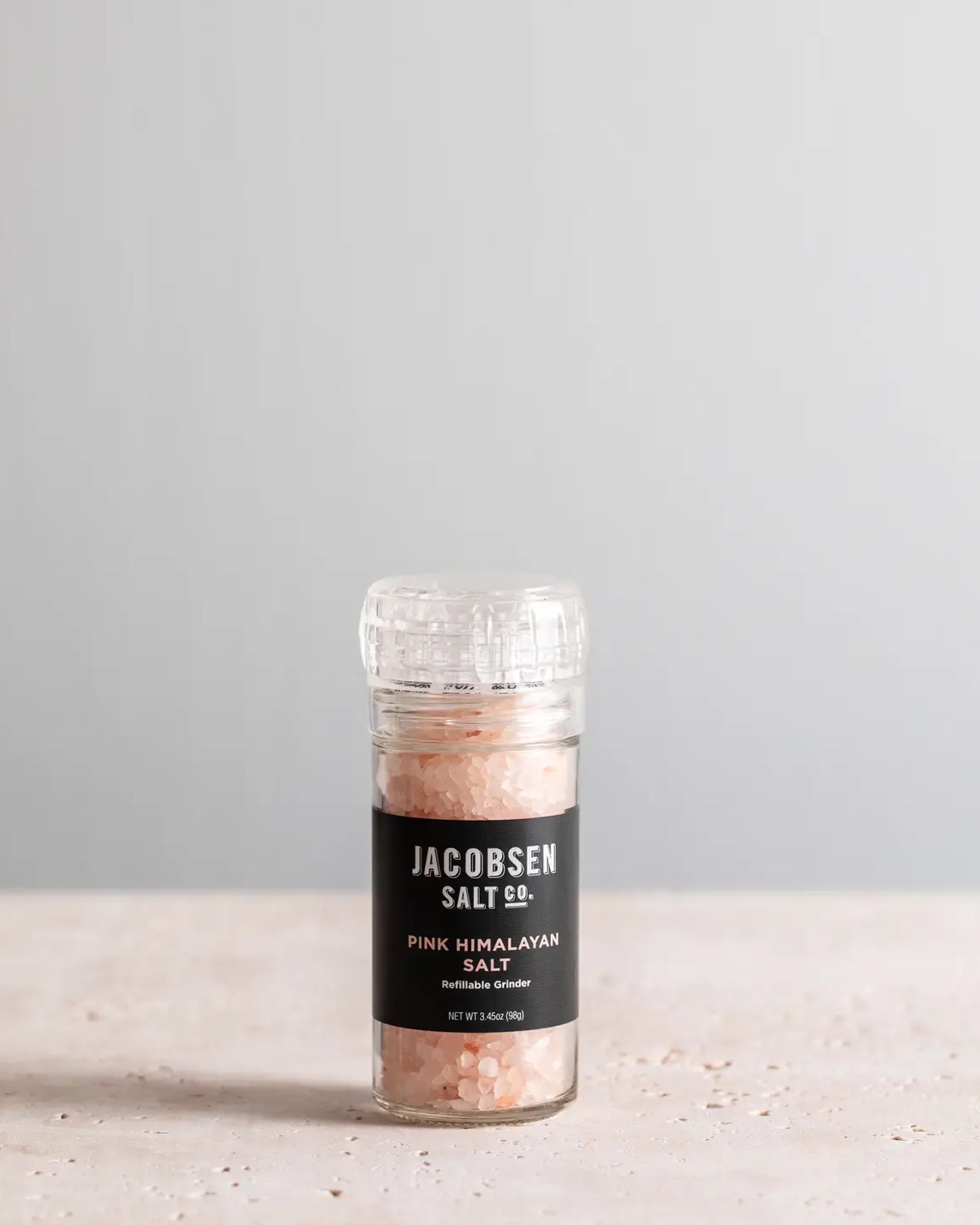 A clear, refillable glass Pink Himalayan Salt Grinder from Faire, filled with mineral-rich salt crystals, sits on a light stone surface against a plain gray background.
