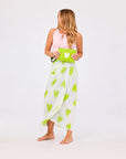 A woman with long, wavy hair wears a pink tank top and a white wrap skirt with green heart prints. She holds the Kerri Rosenthal KR Heart Wristlet and stands barefoot against a plain white background.