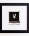 A framed artwork featuring a small, square piece of black paper with a silver Playboy bunny logo in the center. The black paper appears to be attached to a white background, and the entire piece is enclosed in a simple, black wooden frame with clear acrylic for protection, called Art Square Blk Frame by Match South.