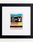 A framed black and white matchbook with "The Playboy Club" and the iconic Playboy bunny logo on it. The matchbook has colored stripes in blue, yellow, orange, and red at the bottom. The clear acrylic frame of the Art Square Blk Frame by Match South hangs elegantly on a plain white wall.