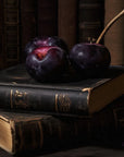An ADLAN Candle by ADLAN Fragrances sits beside two ripe plums with a branch on stacked leather-bound books; violet petals are scattered nearby, and antique books blur in the background.