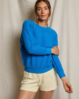 A woman stands before a beige fabric backdrop, wearing the Perfectwhitetee Ziggy Reverse LS Sweatshirt in bright blue and light drawstring shorts, one arm crossed over her body and a relaxed expression.