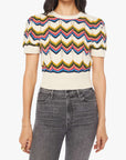 A person wears The Powder Puff knit top by Mother, featuring cream color, multicolored zigzag patterns, and puffed sleeves, styled with high-waisted black jeans. Their head is not visible.