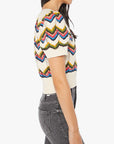 A woman is shown in profile against a white background wearing Mother’s The Powder Puff—high-waisted gray jeans—and a cropped, multicolored chevron-patterned knit top with short sleeves.