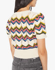 A woman with long dark hair is shown from the back wearing The Powder Puff by Mother, a cropped short-sleeve knit top with colorful zigzag patterns, paired with high-waisted gray jeans.