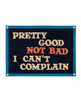 The Oxford Pennant "Pretty Good Not Bad I Can't Complain" banner is dark blue felt with cream and orange text, inspired by John Prine’s “That’s the Way the World Goes Round,” shown on a white background.