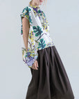 A person with blond hair wears a vibrant floral blouse and Psophia's Air Drift Wide-Leg Culotte with Drawstring Waist. White sandals finish the look as they stand against a plain light backdrop.