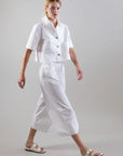 A person in a white short-sleeve button-up shirt, Psophia’s Bouton & Brume Elastic-Waist Cotton Palazzo Pants, and white sandals walks confidently against a gray background, showcasing the pants' flowing silhouette.