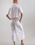 A person faces away, wearing Psophia's Bouton & Brume Short-Sleeve Cotton Bolero over a loose white shirt, with matching wide-leg cropped pants and white slide sandals against a plain light background.