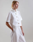 A woman stands in a white studio wearing the Psophia Bouton & Brume Short-Sleeve Cotton Bolero over a collared white shirt and loose white pants, her hands in her pockets as she gazes thoughtfully to the side.