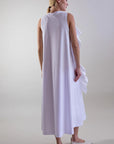 A woman with blonde hair in a low ponytail wears the Psophia Sculptural Rosette Mercerized Cotton Midi Dress in white and matching flat shoes, standing on a light background and facing slightly away from the camera.