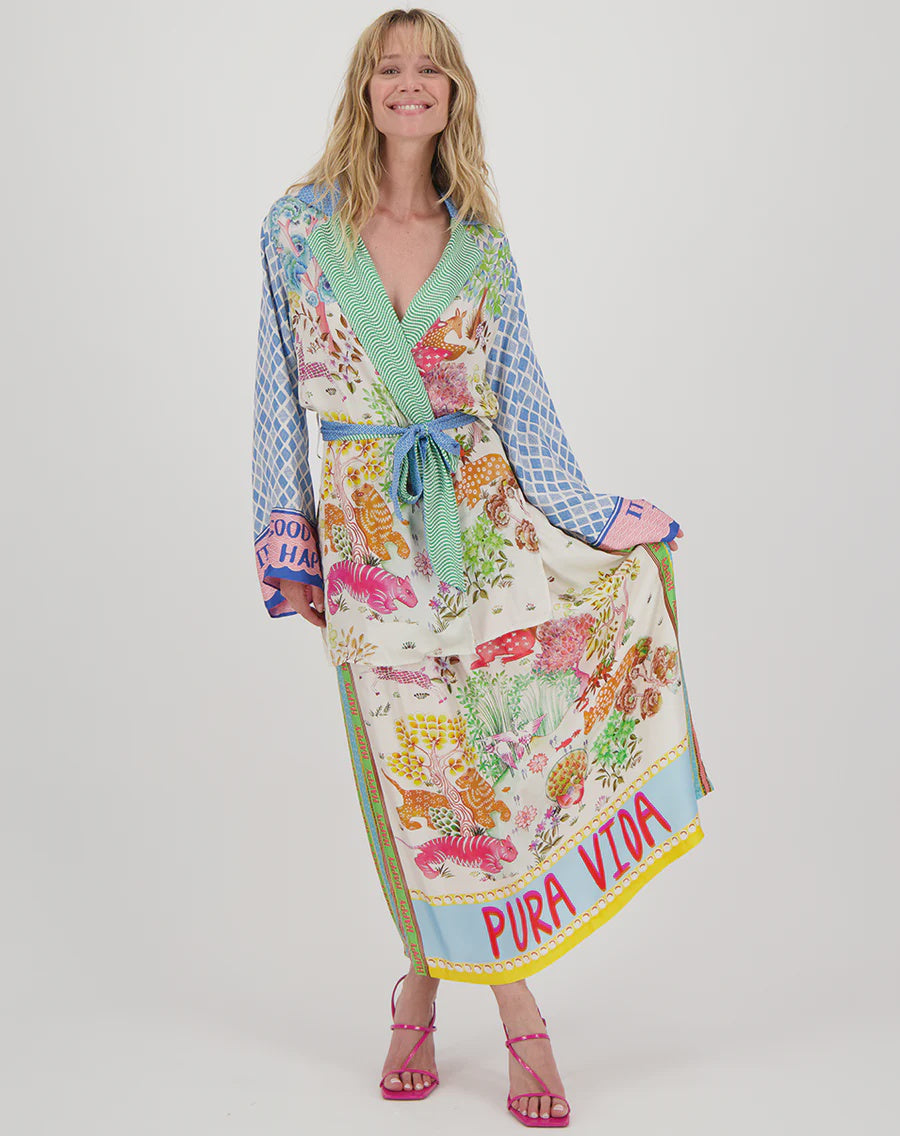 A smiling person wears the Vanessa Midi Skirt by Me369, a colorful floral piece with "PURA VIDA" at the hem. They pair it with pink sandals and radiate joy against a plain white background in their eye-catching ensemble featuring long sleeves and an elastic waist.