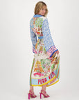 A woman poses with her back slightly turned, holding the hem of a colorful Vanessa Midi Skirt by Me369 featuring various patterns and animals with "PURA VIDA" at the bottom. She accessorizes with pink heels and a matching shirt, smiling slightly against a plain white background.