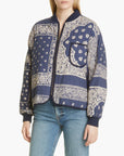A person wearing The Great Inc.'s The Quilted Bomber in navy blue cotton with a cream bandana print, front zip, and curved hem, paired with light blue jeans.