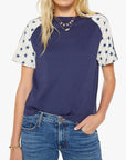 A woman wears The SS Concert Race by Mother, a boxy fit 100% cotton crewneck tee with navy and white star-patterned raglan sleeves, paired with blue jeans and layered necklaces. One hand is in her pocket.