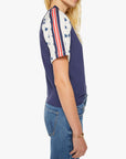 A woman stands in side profile against a plain background, wearing Mother’s The SS Concert Race tee—a short-sleeved, boxy fit navy raglan shirt with white sleeves featuring blue stars and red stripes—paired with blue jeans.