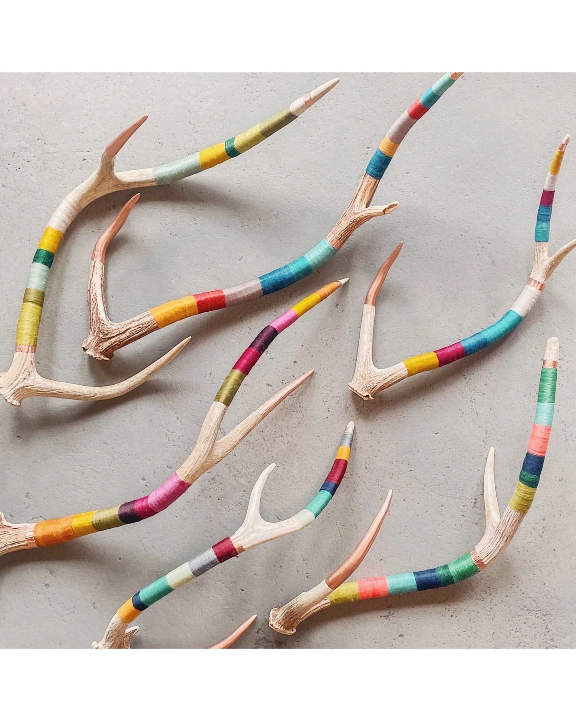 The Faire Wool & Copper wrap Deer Antler features handcrafted antlers on a light grey surface, each wrapped in vibrant colored thread—yellow, blue, red, green, and pink—making them unique decor pieces for any space.