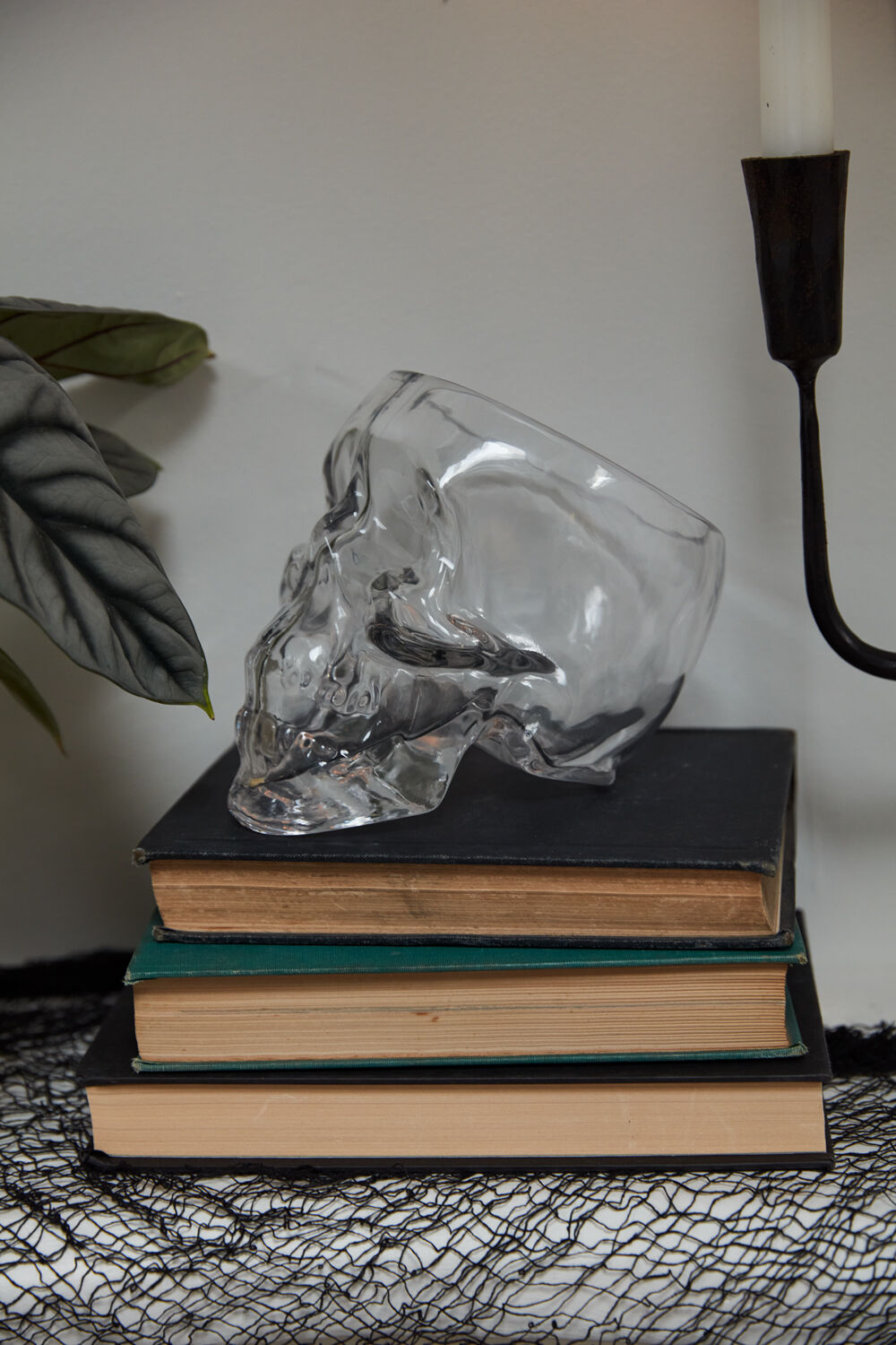 The Accent Decor Reaper Pot, a clear skull-shaped terrarium, sits atop three dark books with a black candle holder and leafy plant, bringing witchy decor vibes to your space.