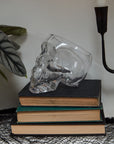 The Accent Decor Reaper Pot, a clear skull-shaped terrarium, sits atop three dark books with a black candle holder and leafy plant, bringing witchy decor vibes to your space.