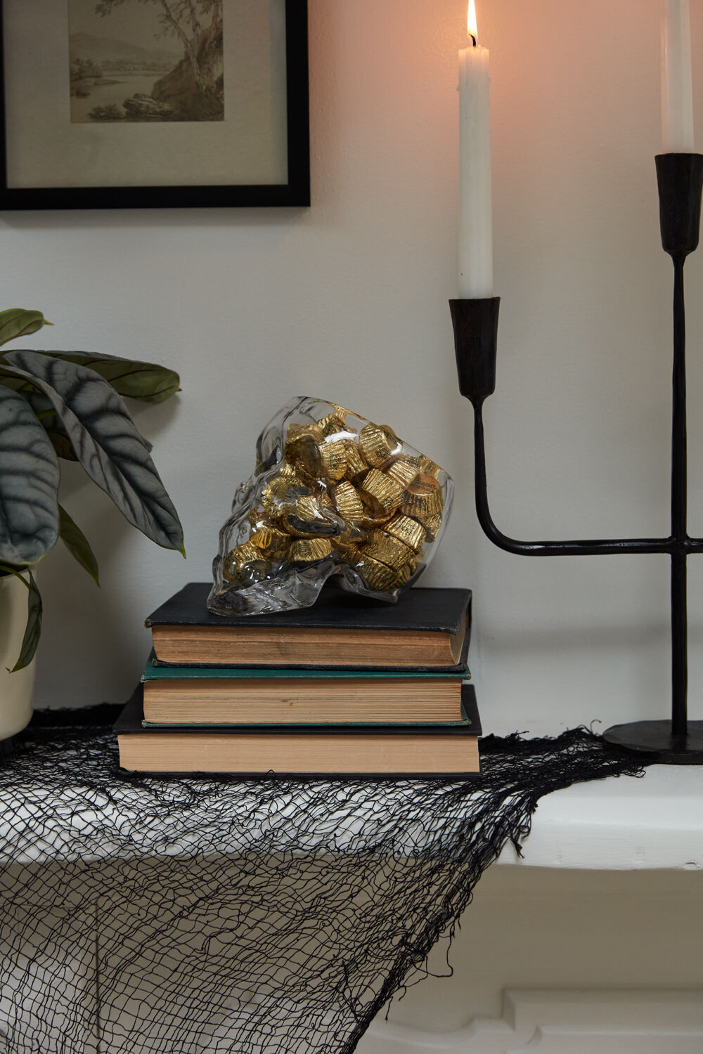 Accent Decor’s Reaper Pot, a silver skull planter filled with gold-wrapped chocolates, sits atop books and lace beside a black candleholder, white candles, and leafy plant—ideal for witchy decor or a spooky planter display.