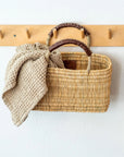 The Gardener Long Basket by Faire, handcrafted in Morocco from woven palm leaves with brown leather handles, hangs on a wooden peg rack with a textured beige cloth draped over the side against a white wall.