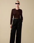 Wearing black sunglasses and the Sold Out NYC Refined Crew eco-friendly dark brown Merino wool sweater, tucked into high-waisted black wide-leg trousers with a black belt, against a beige background.