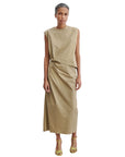 A person with gray hair styled back wears the Rachel Comey Rini Dress, a modern elegant sleeveless tan maxi with a draped waist, paired with yellow strappy heels, standing against a plain white background.